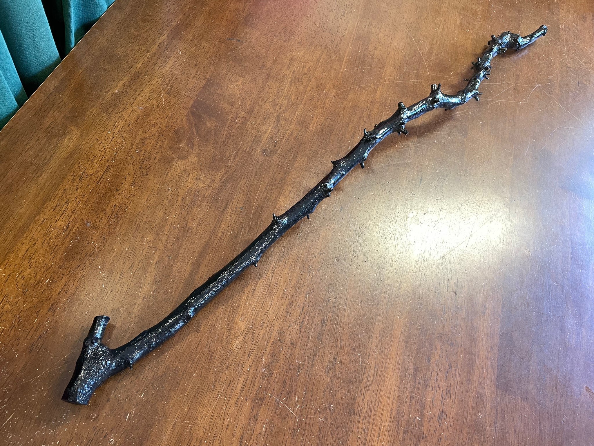 A long, dark blackthorn walking stick laid on a polished wood table, the trunk showing the trimmed nubs of former thorns along its length and a small T-shaped handle at the top.