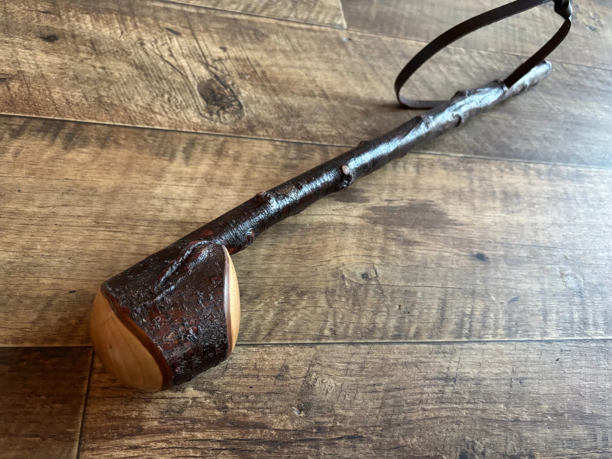 A finished blackthorn shillelagh laid on a wood floor, with a dark stained root knob, a polished shaft, and a leather wrist strap looped at the head.