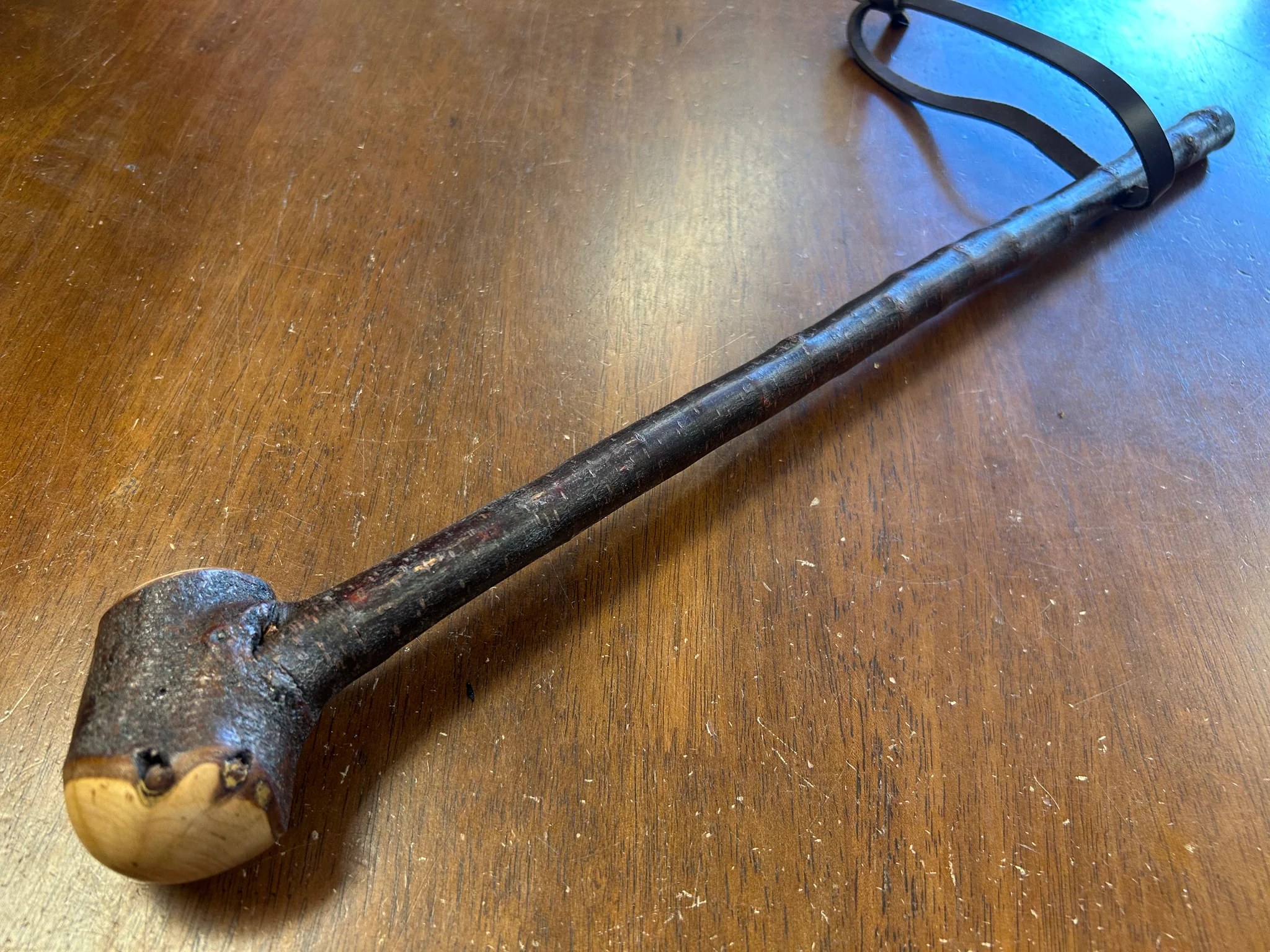 A short blackthorn shillelagh laid horizontally on a wood floor next to a coiled leather wrist strap, with a heavy dark root knob at one end and the polished shaft tapering toward the foot.