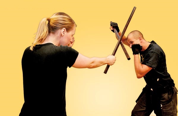 A photograph of two practitioners of eskrima in mid-exchange, each holding a single rattan stick, captured at a moment of contact between the two sticks at chest height.